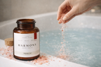 Hand pouring Himalayan luxury pink salt from a bottle labeled 'Harmony' into a bathtub.